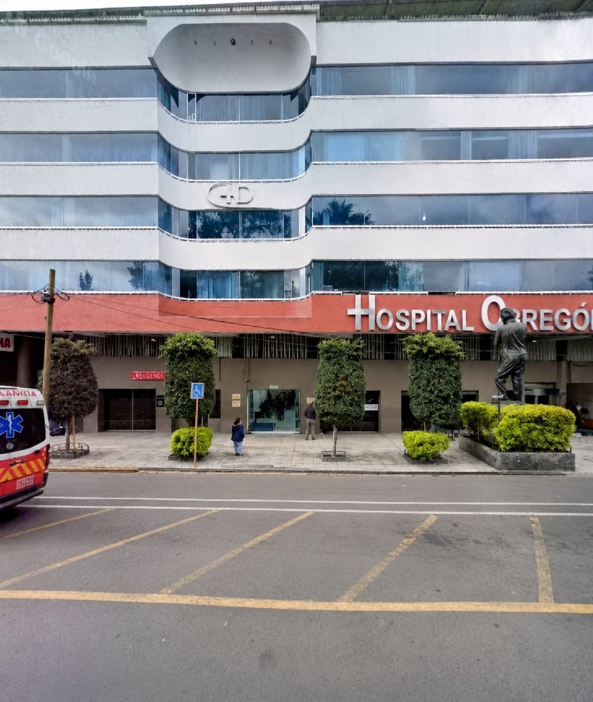 Hospital Grego building exterior with white and red striped facade, curved balconies, and ground-level entrance with landscaping
